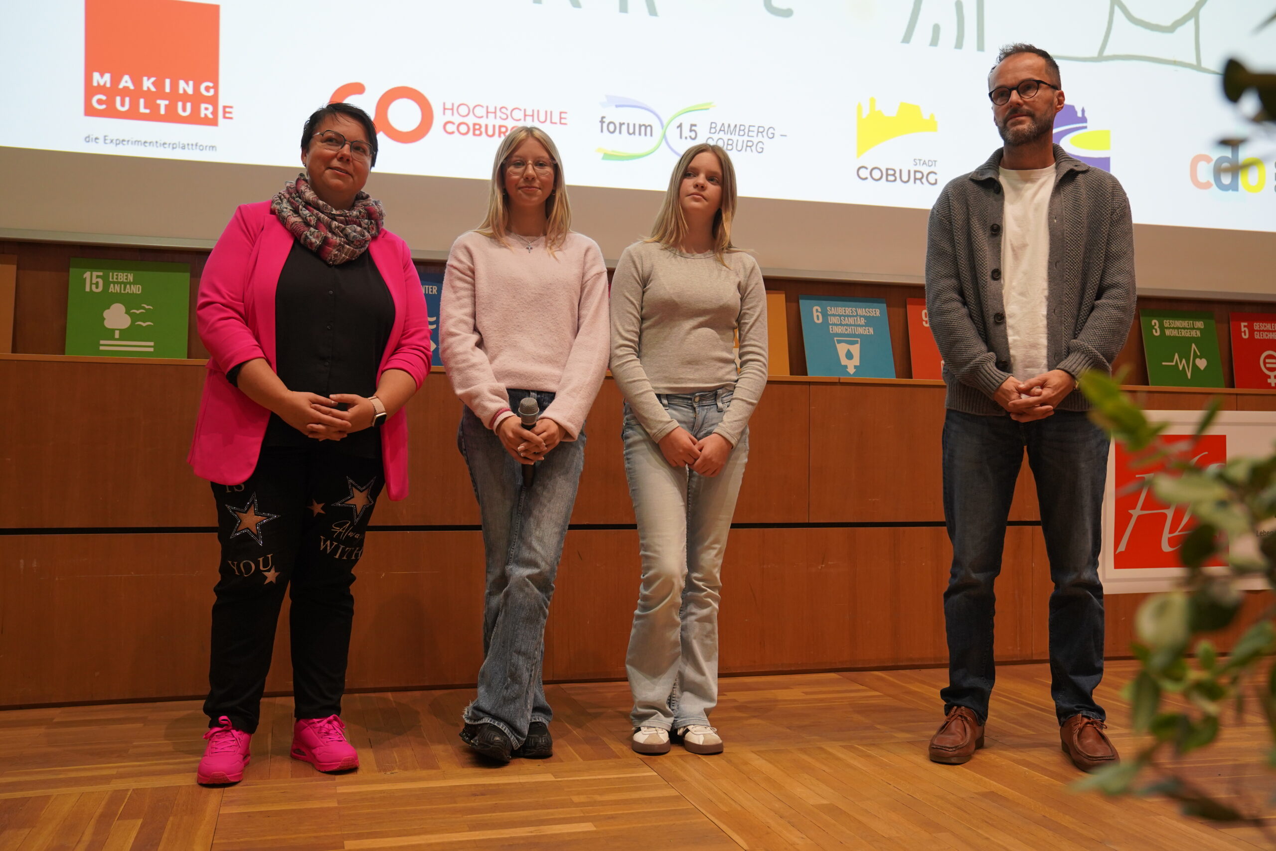Zwei Frauen und zwei Männer stehen auf einer Bühne vor einer Projektionswand für ein automatisch aufgenommenes Gruppenfoto. at Hochschule Coburg