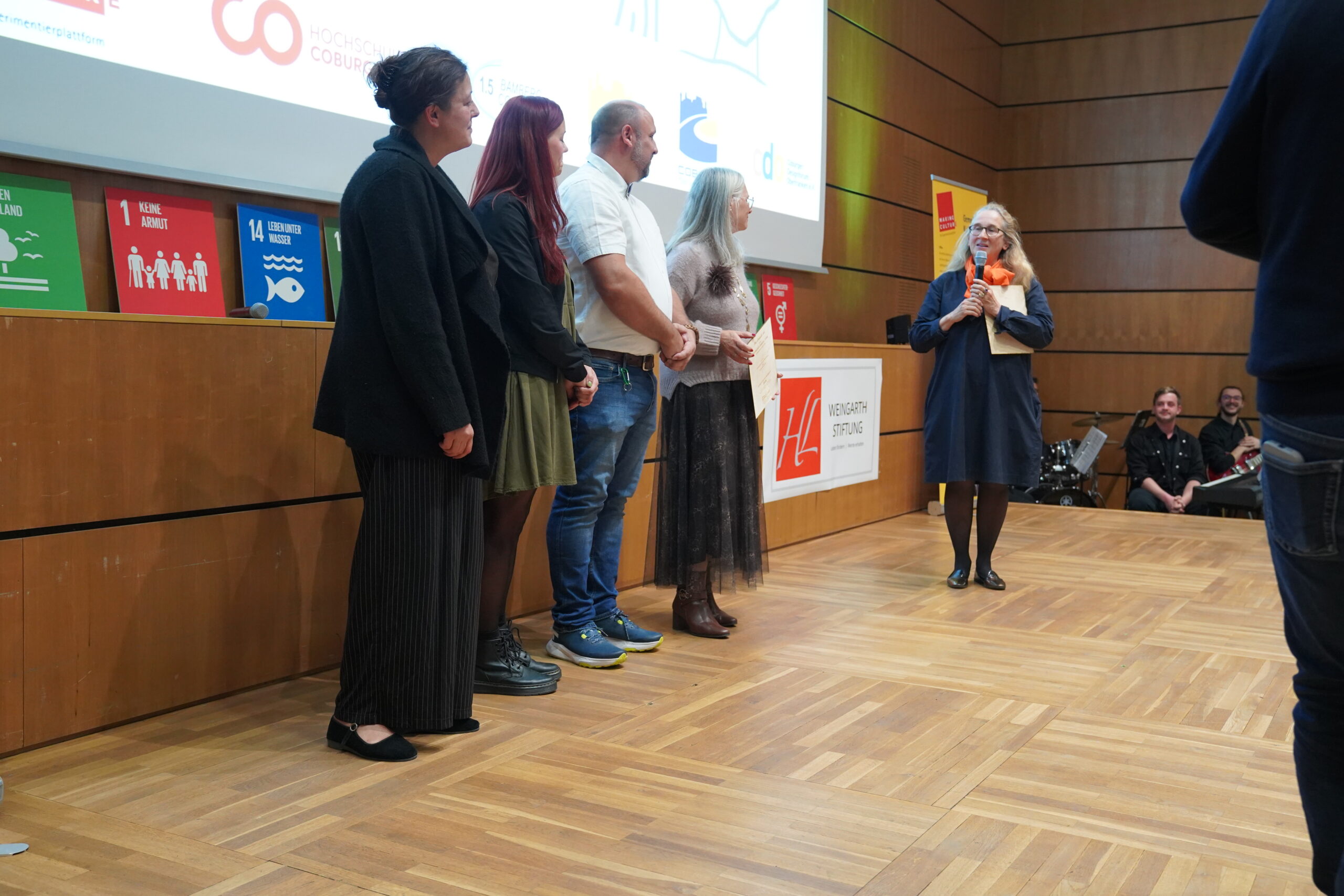 Eine Frau spricht auf einer Bühne in ein Mikrofon, während vier weitere Personen während einer Präsentation zum Thema „Automatisierung“ in einer Reihe neben ihr stehen. at Hochschule Coburg