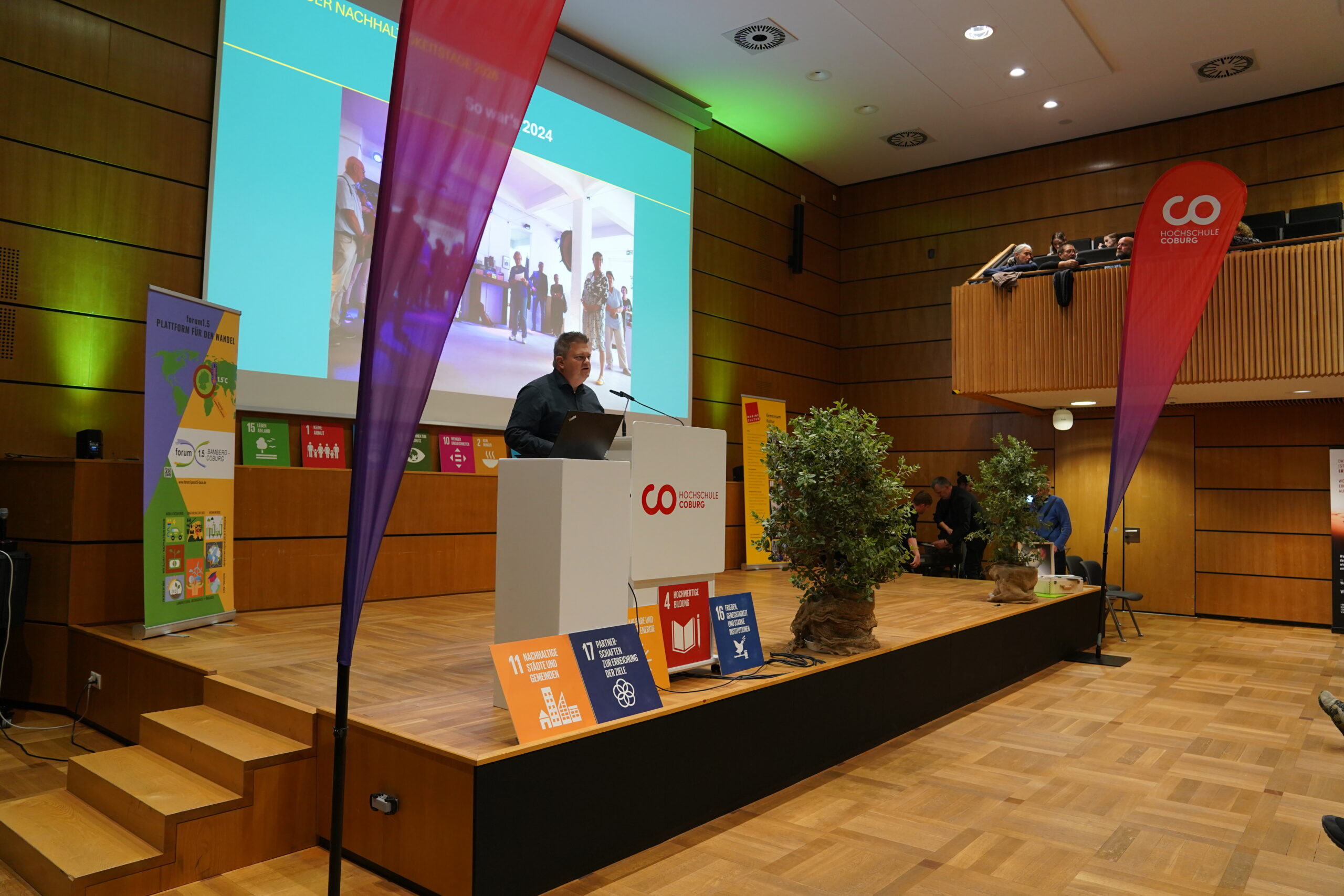 Ein Mann spricht während einer Präsentation zum Thema „Automatischer Entwurf“ an der Hochschule Coburg von einem Podium aus, während hinter ihm Projektbilder auf einer großen Leinwand gezeigt werden. at Hochschule Coburg