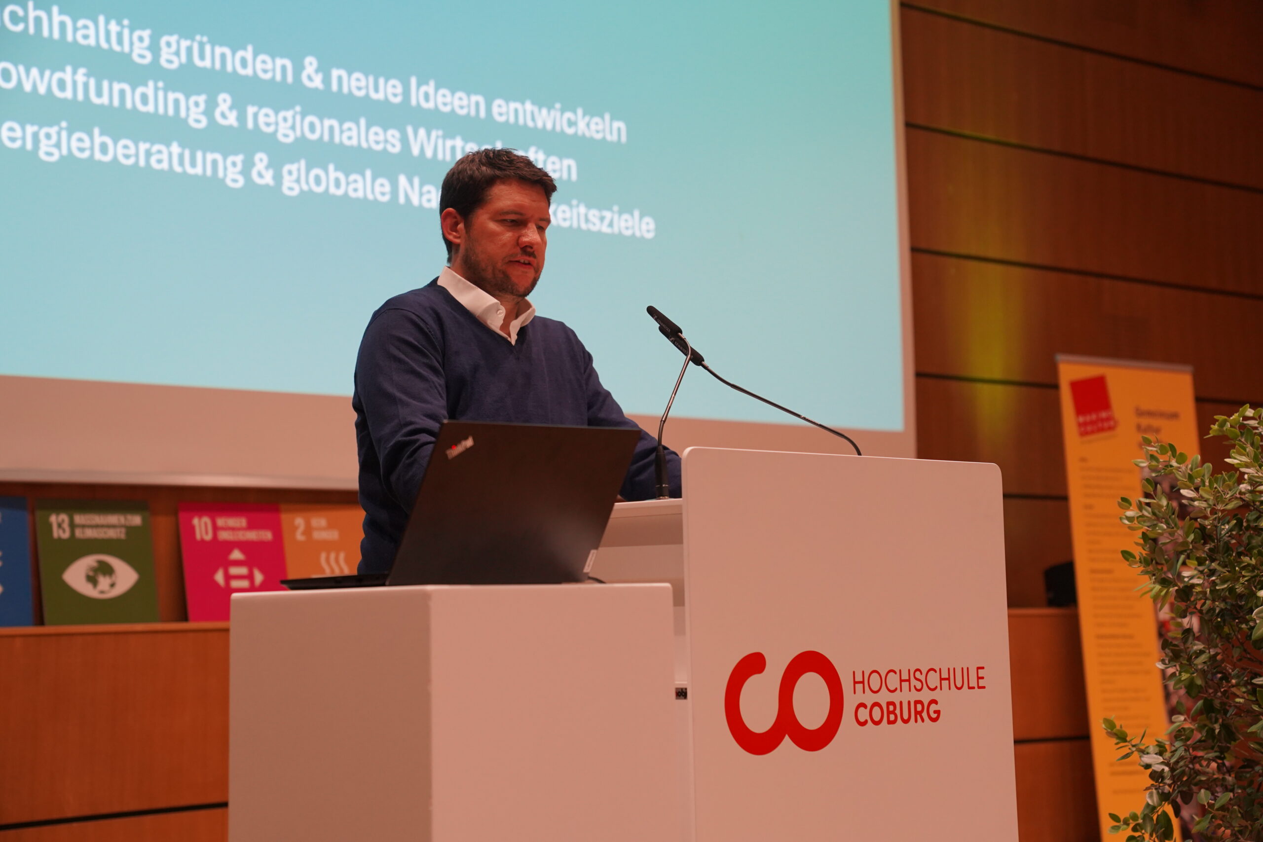 Ein Mann spricht auf einem Podium der Hochschule Coburg vor einer Präsentationsleinwand über nachhaltige Entwicklung und automatisiertes Design. at Hochschule Coburg