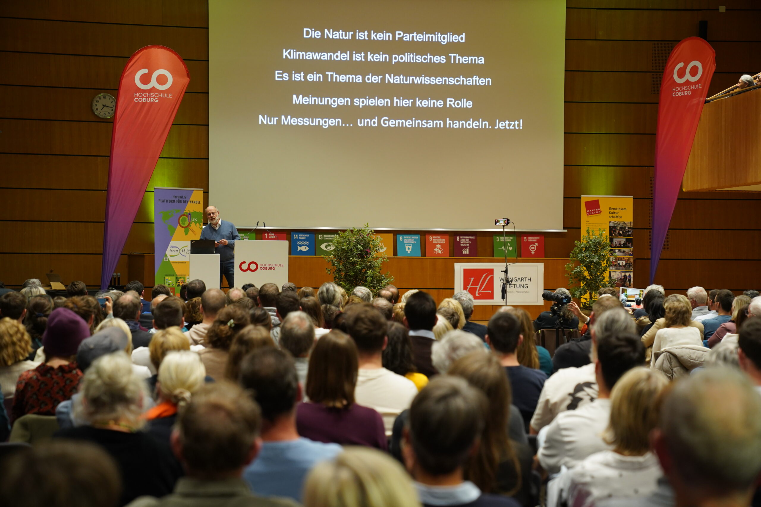 Ein Redner steht an einem Rednerpult vor dem Publikum in einem Hörsaal, während auf einer Leinwand ein „Automatischer Entwurf“ zum Thema Natur und Klimawandel gezeigt wird. at Hochschule Coburg
