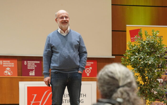 Ein Mann in einem blauen Pullover und Jeans steht auf einer Bühne und spricht vor Publikum über automatisiertes Design. at Hochschule Coburg