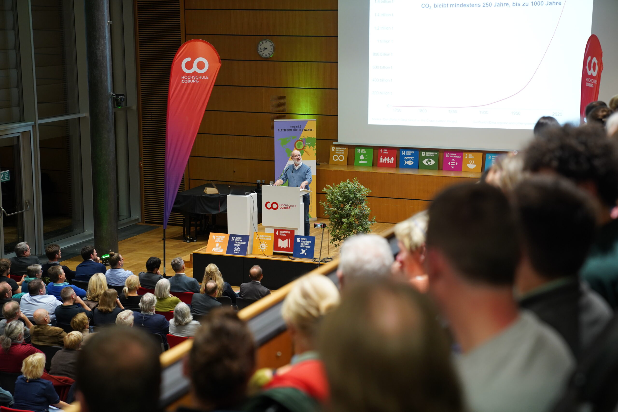 Ein Redner steht auf einem Podium und hält in einem großen Hörsaal einen Vortrag, während hinter ihm auf einer Leinwand eine Grafik zu den CO₂-Emissionen sowie Symbole für die Ziele für nachhaltige Entwicklung zu sehen sind. at Hochschule Coburg