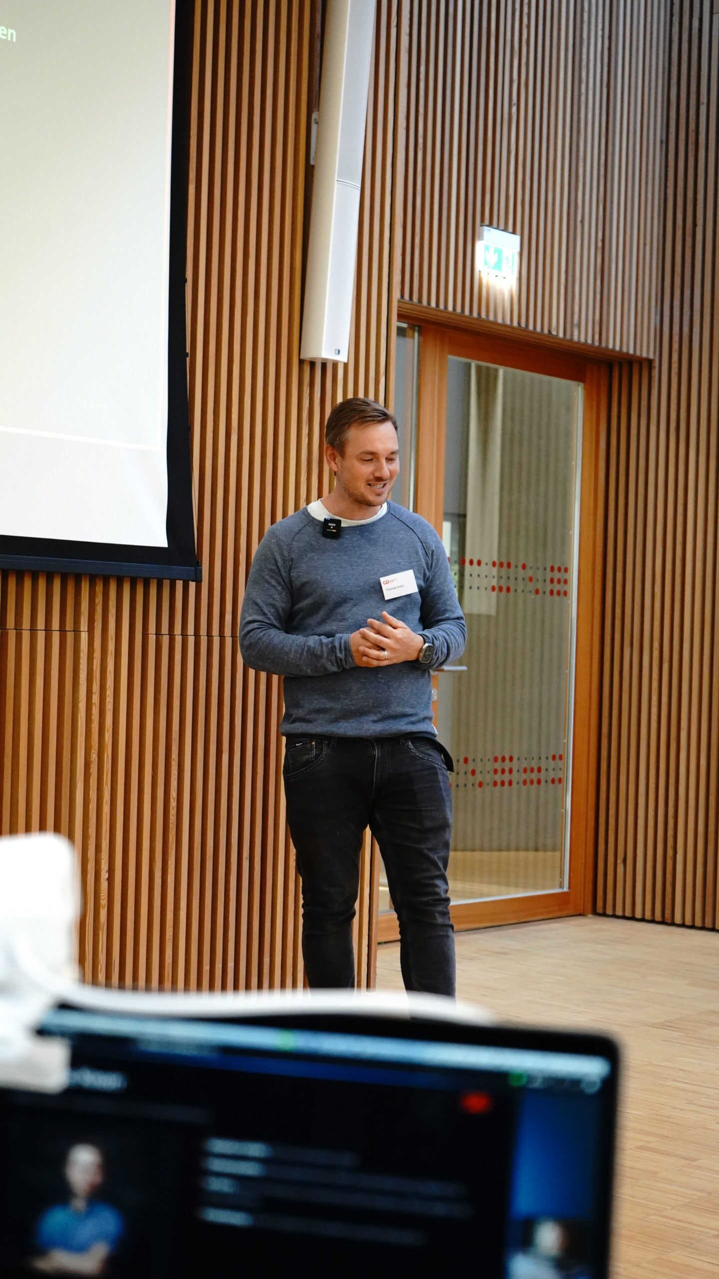 Ein KI-Lösungsarchitekt in einem blauen Pullover steht vor einer holzvertäfelten Wand und hält eine Präsentation. at Hochschule Coburg