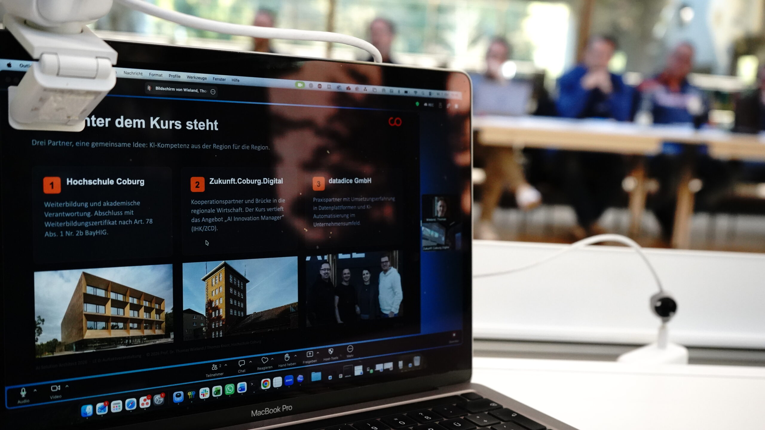 Auf dem Bildschirm eines Laptops wird eine Präsentation zur Entwicklung von KI-Lösungen mit dem Titel „Drei Partner, eine gemeinsame Idee“ für ein gemeinsames KI-Projekt angezeigt, wobei im Hintergrund unscharf abgebildete Teilnehmer zu sehen sind. at Hochschule Coburg