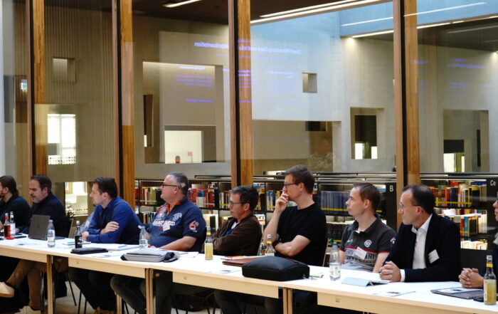 Eine Reihe von Personen, die während eines Treffens der KI-Lösungsarchitekten in einer Bibliothek an einem langen Tisch sitzen. at Hochschule Coburg