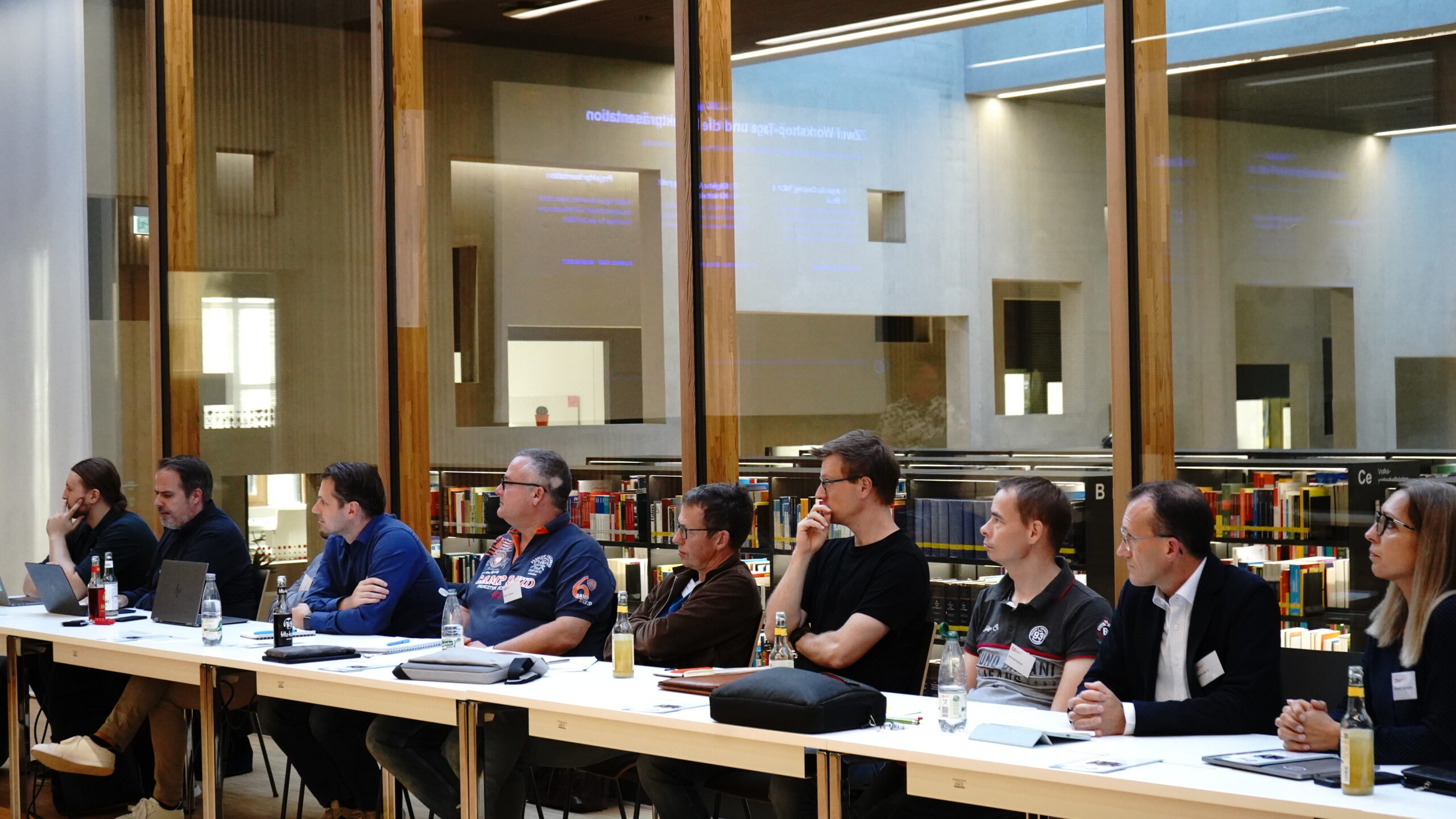 Eine Reihe von Personen, die während eines Treffens der KI-Lösungsarchitekten in einer Bibliothek an einem langen Tisch sitzen. at Hochschule Coburg
