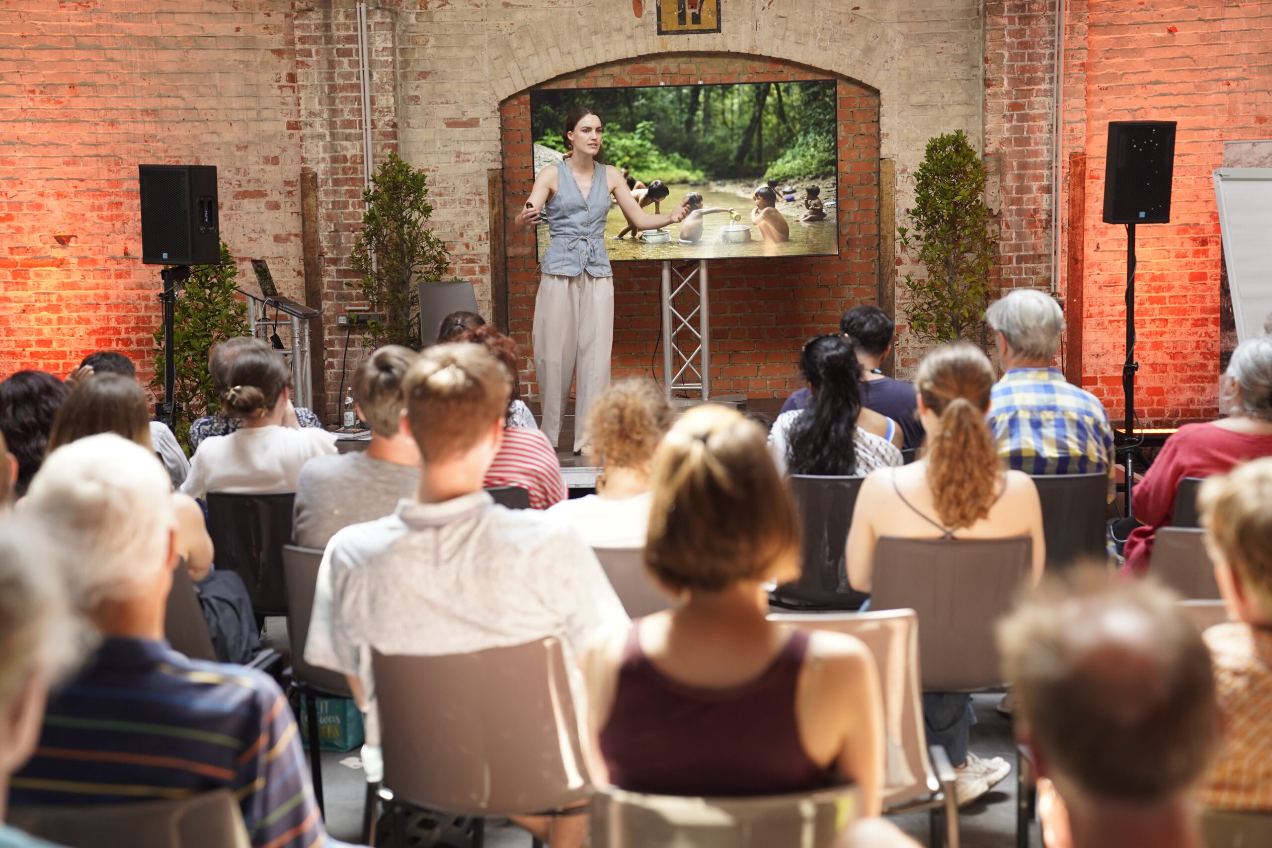 Eine Frau hält auf der Bühne vor einem Publikum einen Vortrag vor einer Leinwand, auf der Menschen beim Baden in einem Fluss zu sehen sind, und hebt dabei die Themen Lebensqualität und Nachhaltigkeit hervor. at Hochschule Coburg