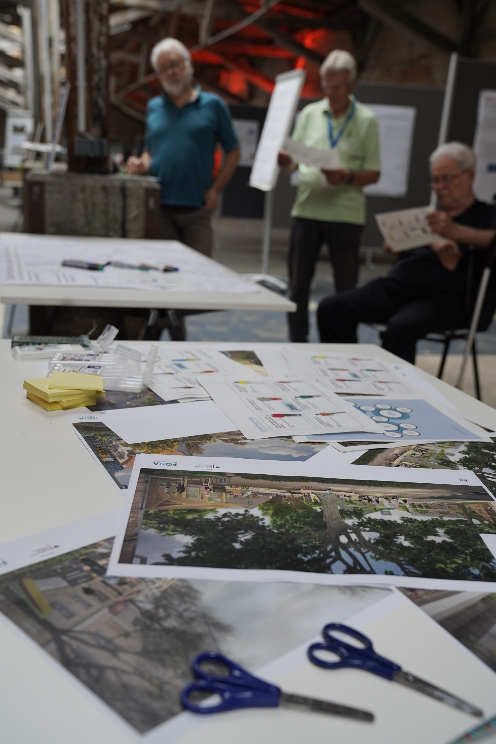 Nahaufnahme eines Tisches, auf dem Architekturzeichnungen und Haftnotizen verstreut liegen, im Hintergrund sind drei Männer zu sehen, die Unterlagen für die Nachhaltigkeitstage durchsehen. at Hochschule Coburg
