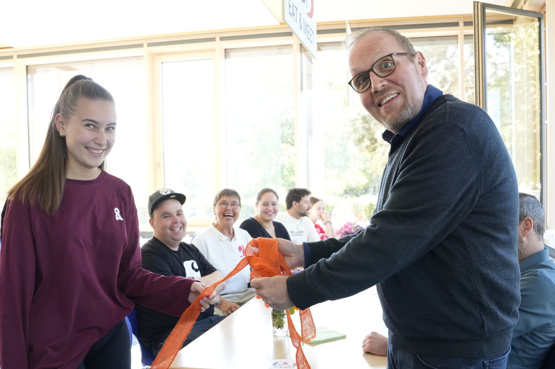 Ein Mann und eine junge Frau halten gemeinsam ein orangefarbenes Band in der Hand und lächeln in die Kamera, während hinter ihnen in einem Raum weitere Personen an Tischen sitzen. at Hochschule Coburg