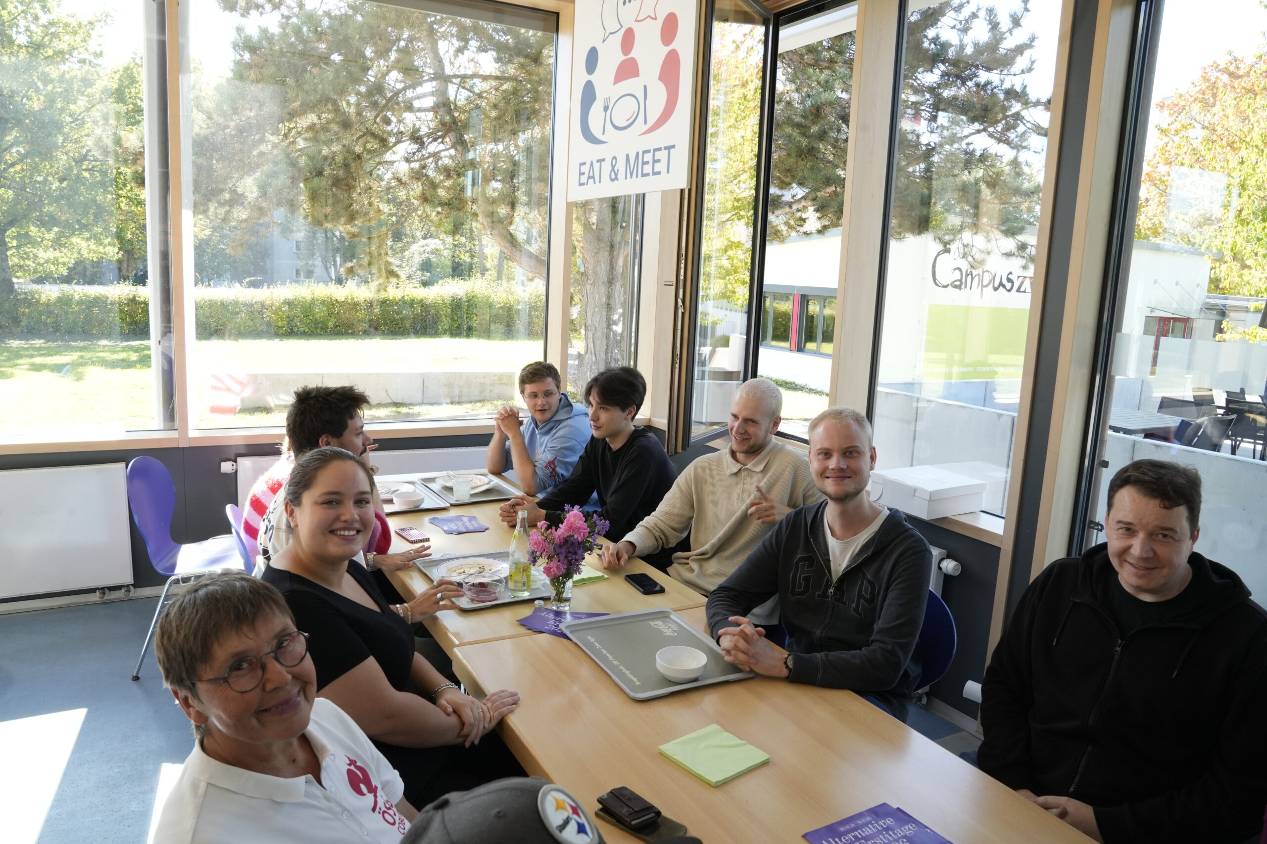 Eine Gruppe von sieben Personen sitzt an einem langen Tisch in einem sonnendurchfluteten Speiseraum, an dessen Fenster ein Schild mit der Aufschrift „Eat & Meet“ hängt. at Hochschule Coburg