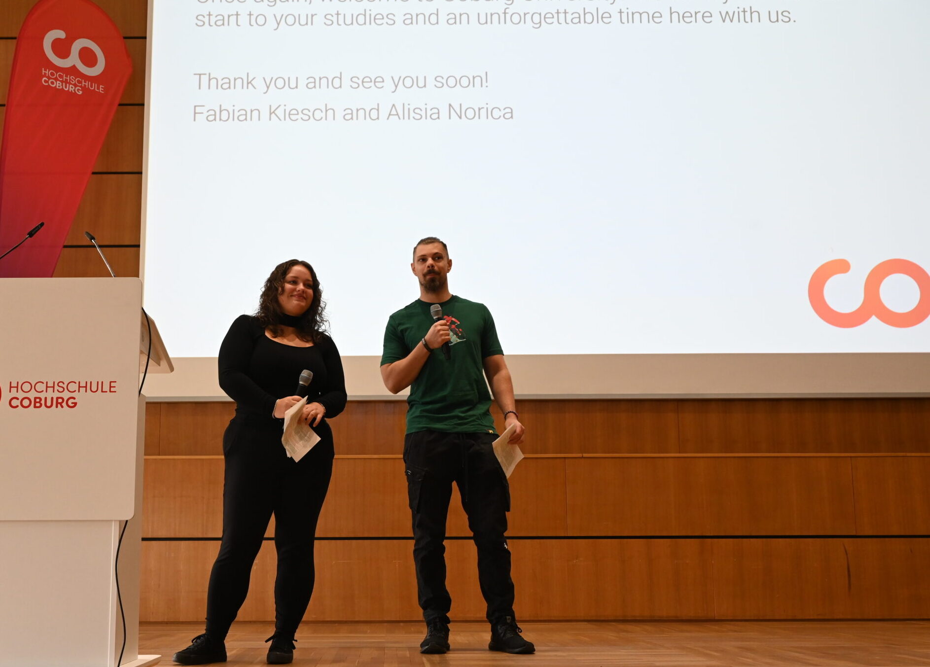 Zwei Personen mit Mikrofonen stehen auf einer Bühne vor einer Leinwand und heißen die Studierenden an der Hochschule Coburg zu ihrem „Studium Coburg“ willkommen. at Hochschule Coburg