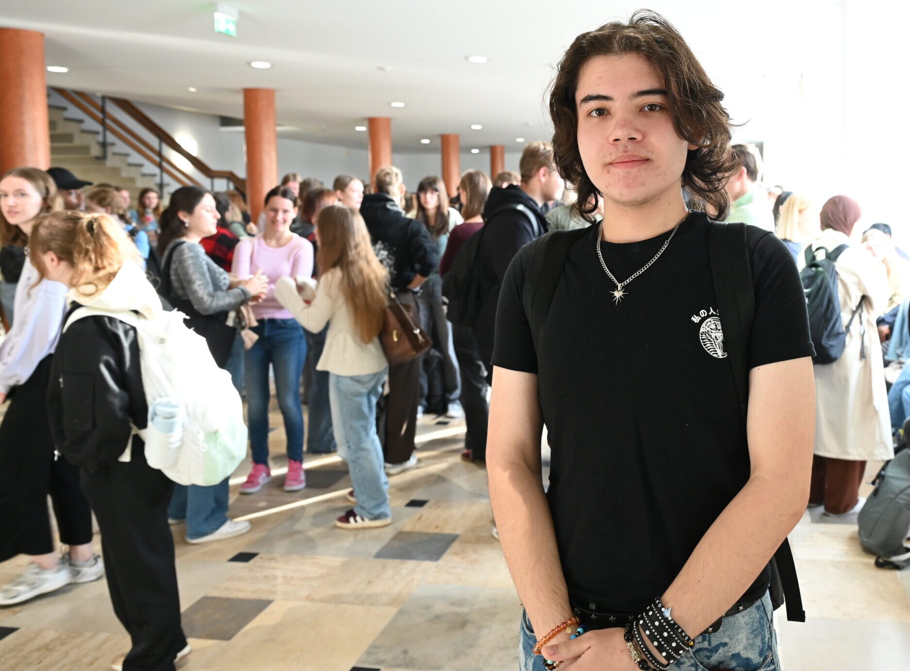 Ein junger Mensch mit schulterlangen Haaren und einem schwarzen T-Shirt steht in einem überfüllten Flur der Hochschule Coburg. at Hochschule Coburg