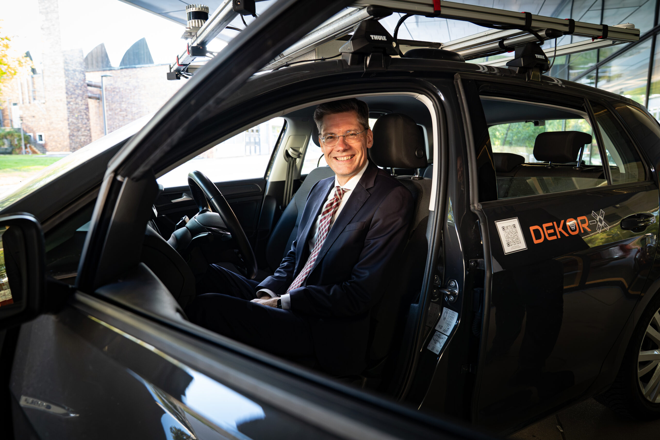 Ein Mann in Anzug und Brille sitzt auf dem Fahrersitz eines „DEKOR“-Autos, das die Zukunft des autonomen Fahrens in ländlichen Gebieten verkörpert. at Hochschule Coburg