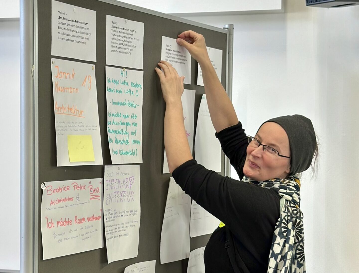 Eine Frau mit Brille und schwarzer Mütze heftet einen Zettel an eine Anschlagtafel bei der „aCOmmen 2026“, die mit handgeschriebenen Schildern für die Fakultät Design + Bauen übersät ist. at Hochschule Coburg