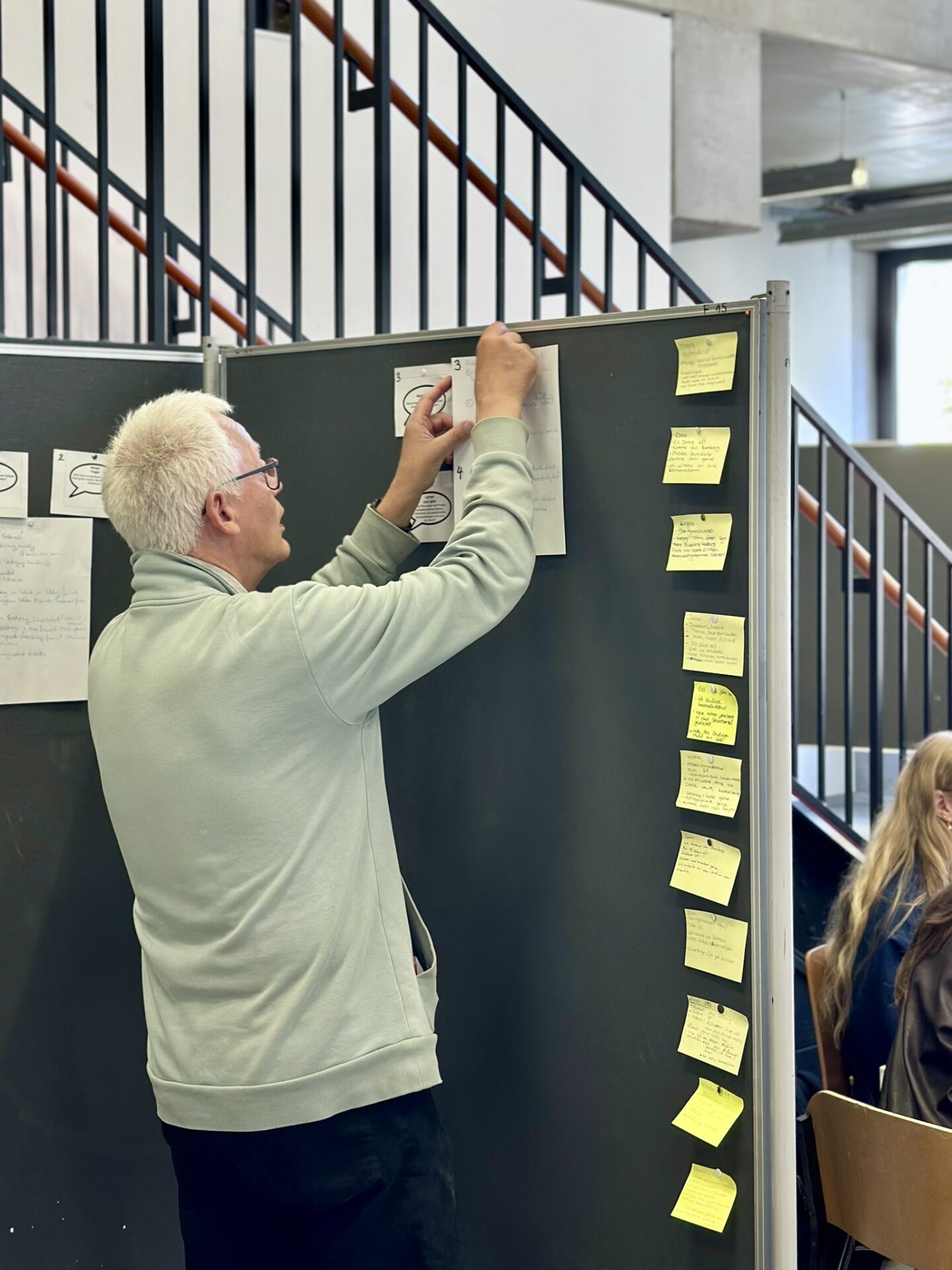 Eine Person heftet ein Blatt Papier an eine mit Haftnotizen bedeckte anCOmmen-2026-Pinnwand. at Hochschule Coburg