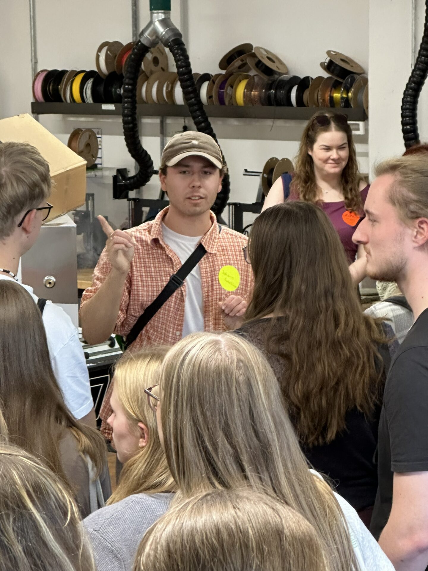 Ein junger Mann in einem karierten Hemd und einer Baseballkappe gestikuliert, während er zu Erstsemestern in einem „Design und Bauen“-Workshop spricht; im Hintergrund stehen Regale mit Filamentspulen. at Hochschule Coburg