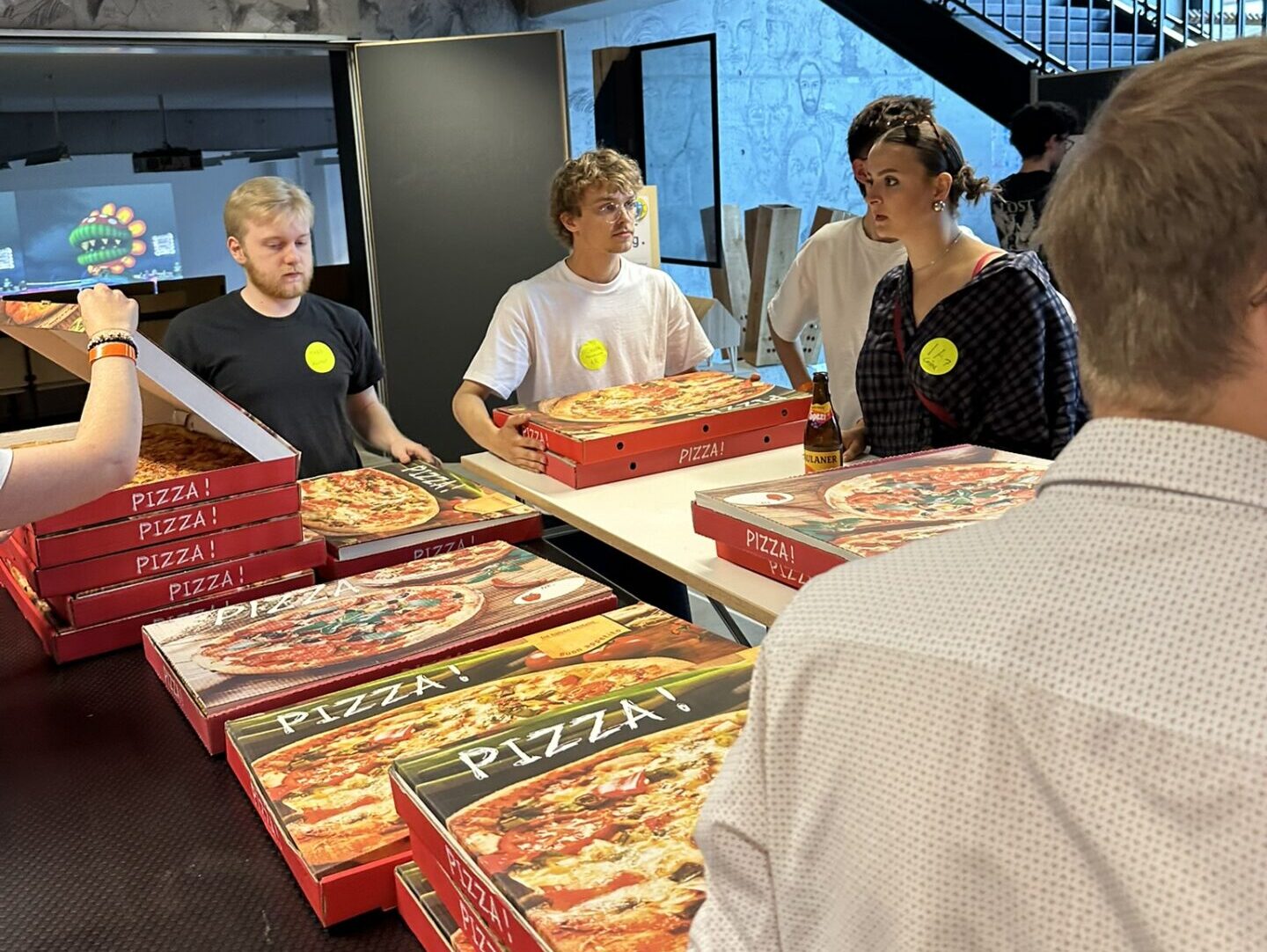 Im Rahmen der Veranstaltung „Design und Bauen“ standen Erstsemester in einem großen Raum um Tische herum, auf denen mehrere Pizzakartons gestapelt waren. at Hochschule Coburg