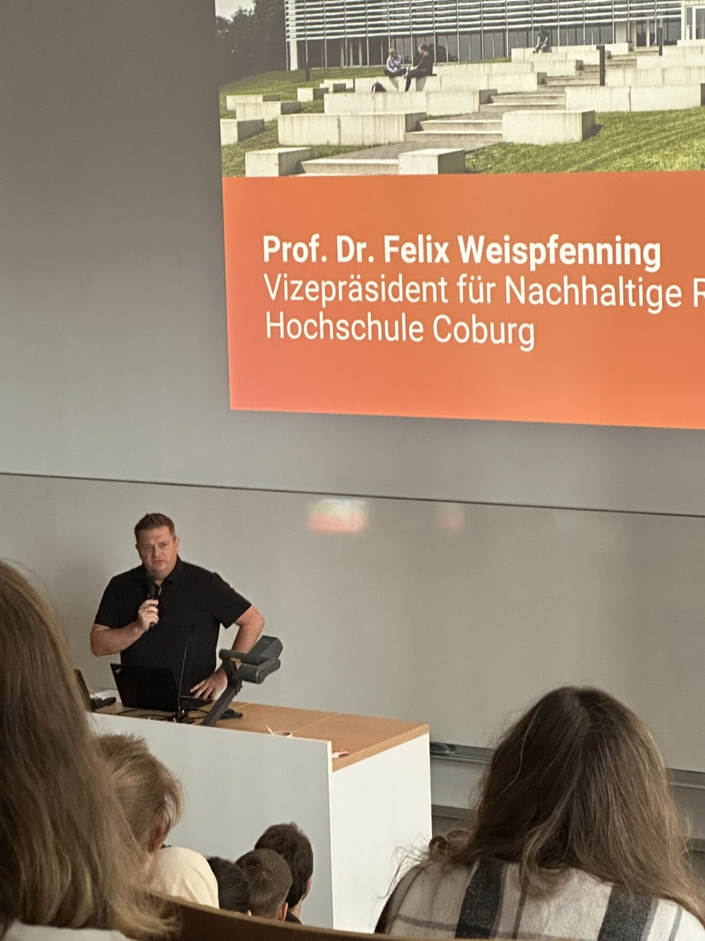Prof. Dr. Felix Weispfenning hält während der Veranstaltung „anCOmmen 2026“ für Studienanfänger eine Rede auf einem Podium, hinter ihm ist auf einer Leinwand seine Rolle im Bereich der nachhaltigen Entwicklung an der Hochschule Coburg dargestellt. at Hochschule Coburg