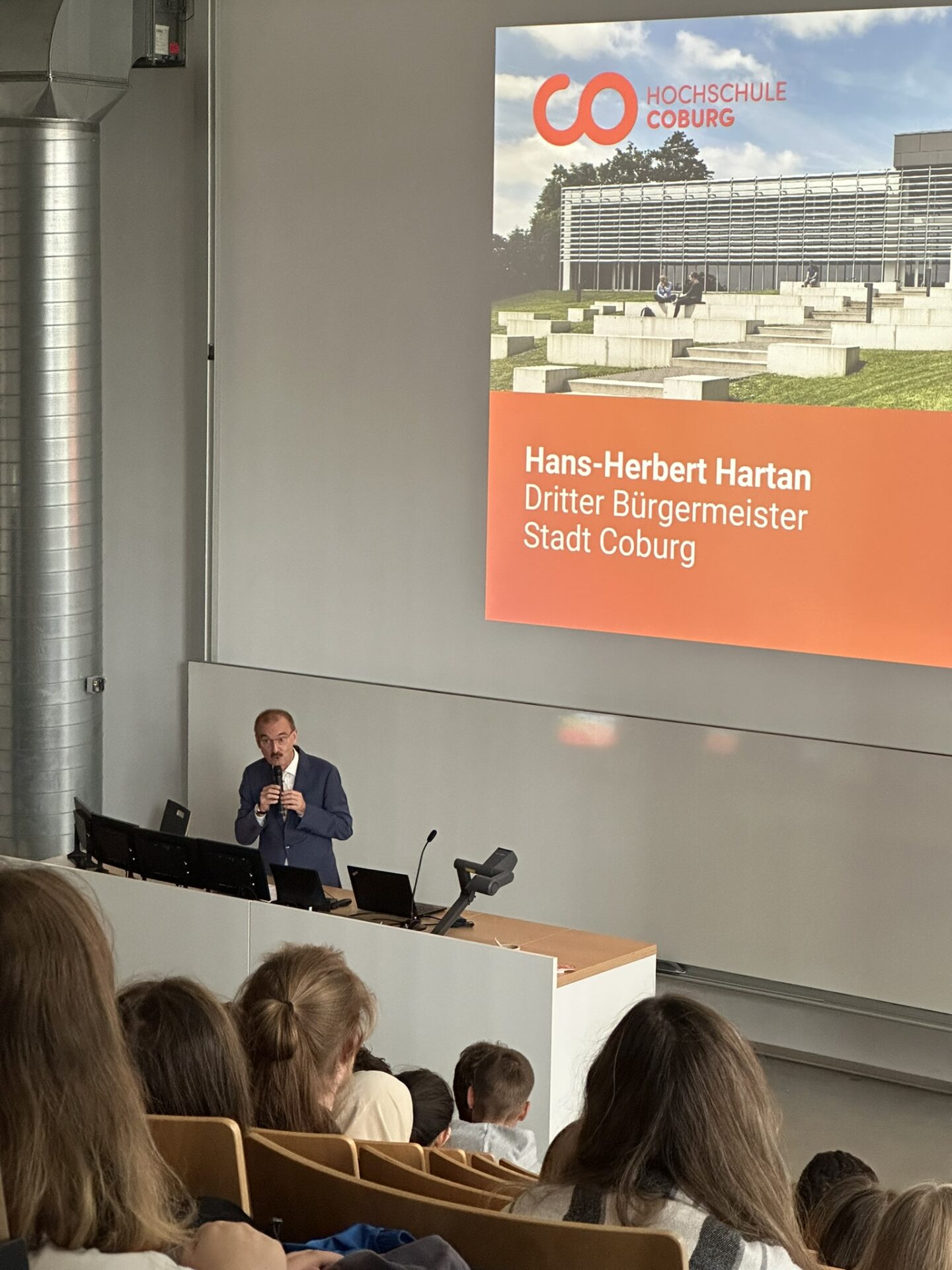 Hans-Herbert Hartan, dritter Bürgermeister der Stadt Coburg, spricht während einer Veranstaltung im Rahmen von „Studieren in Coburg“ an einem Rednerpult in einem Hörsaal, hinter ihm befindet sich eine Leinwand. at Hochschule Coburg