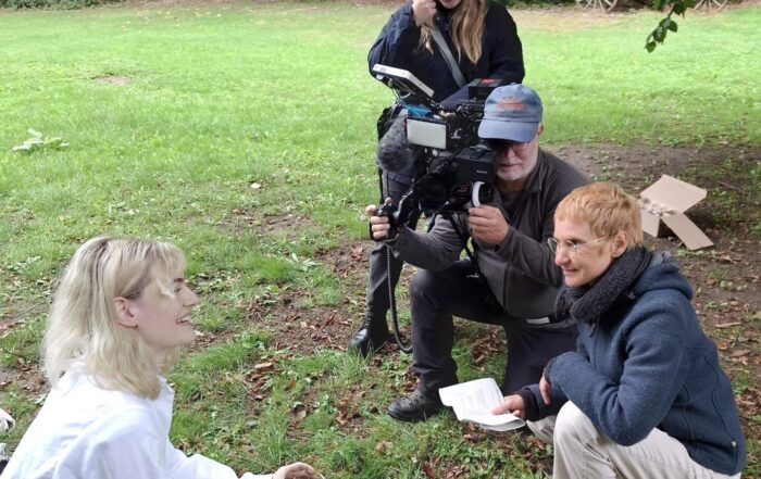 Ein Kameramann, ein Tontechniker und ein Interviewer filmen eine Frau in weißer Kleidung, die mit archäologischen Werkzeugen auf dem Rasen kniet – eine Szene, die die Design-Automatisierung widerspiegelt. at Hochschule Coburg