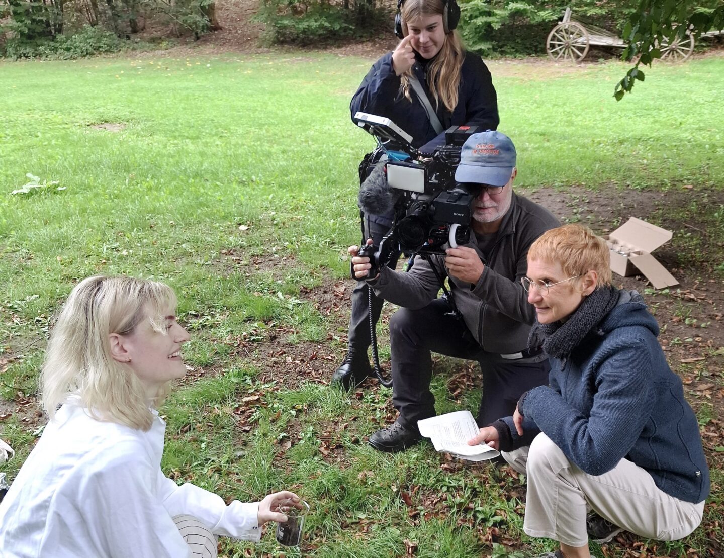 Ein Kameramann, ein Tontechniker und ein Interviewer filmen eine Frau in weißer Kleidung, die mit archäologischen Werkzeugen auf dem Rasen kniet – eine Szene, die die Design-Automatisierung widerspiegelt. at Hochschule Coburg