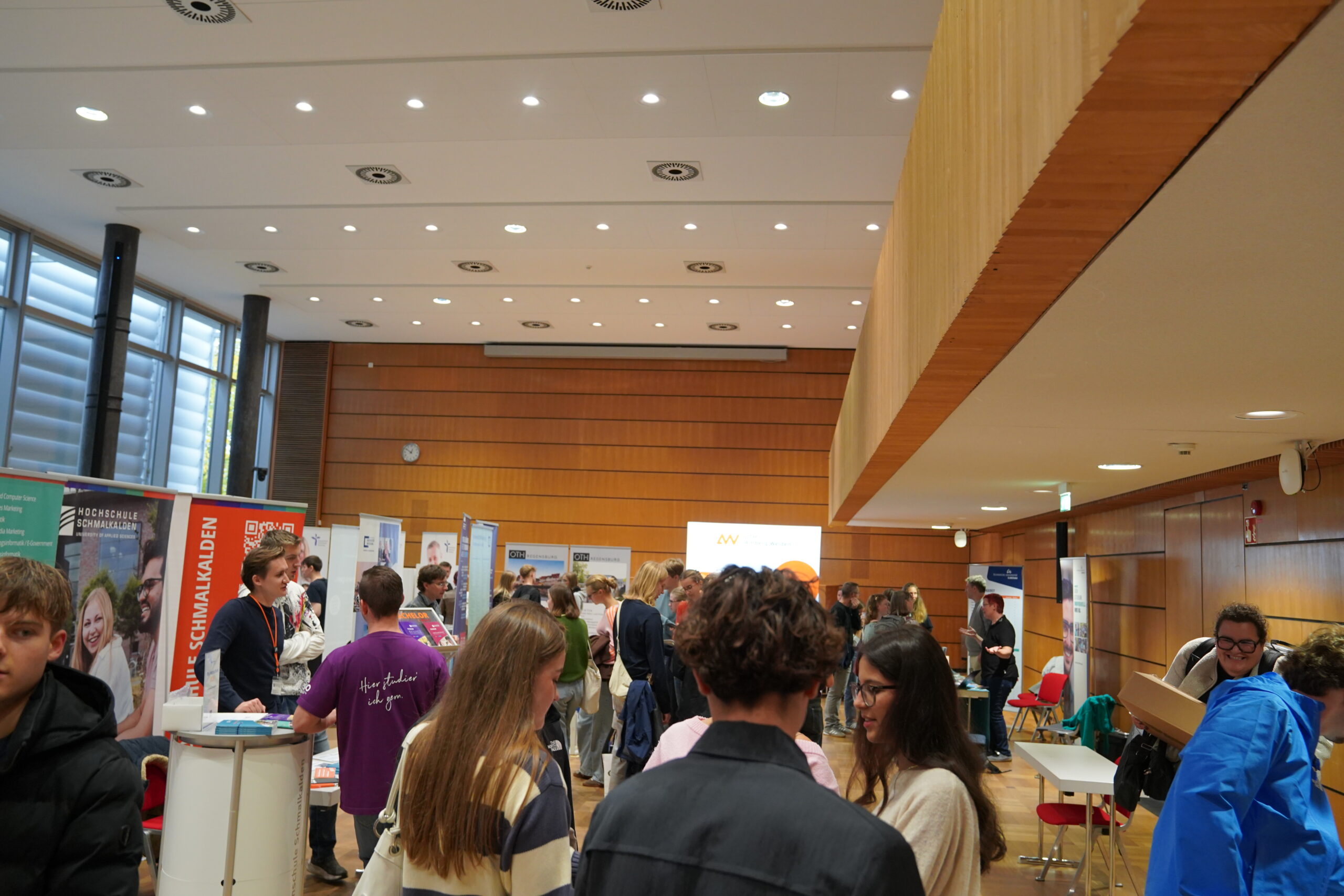 Die Studierenden besuchen die Coburger Studienmesse, eine Hochschulinformationsmesse mit verschiedenen Ständen und Bannern in einer großen, modernen Halle. at Hochschule Coburg
