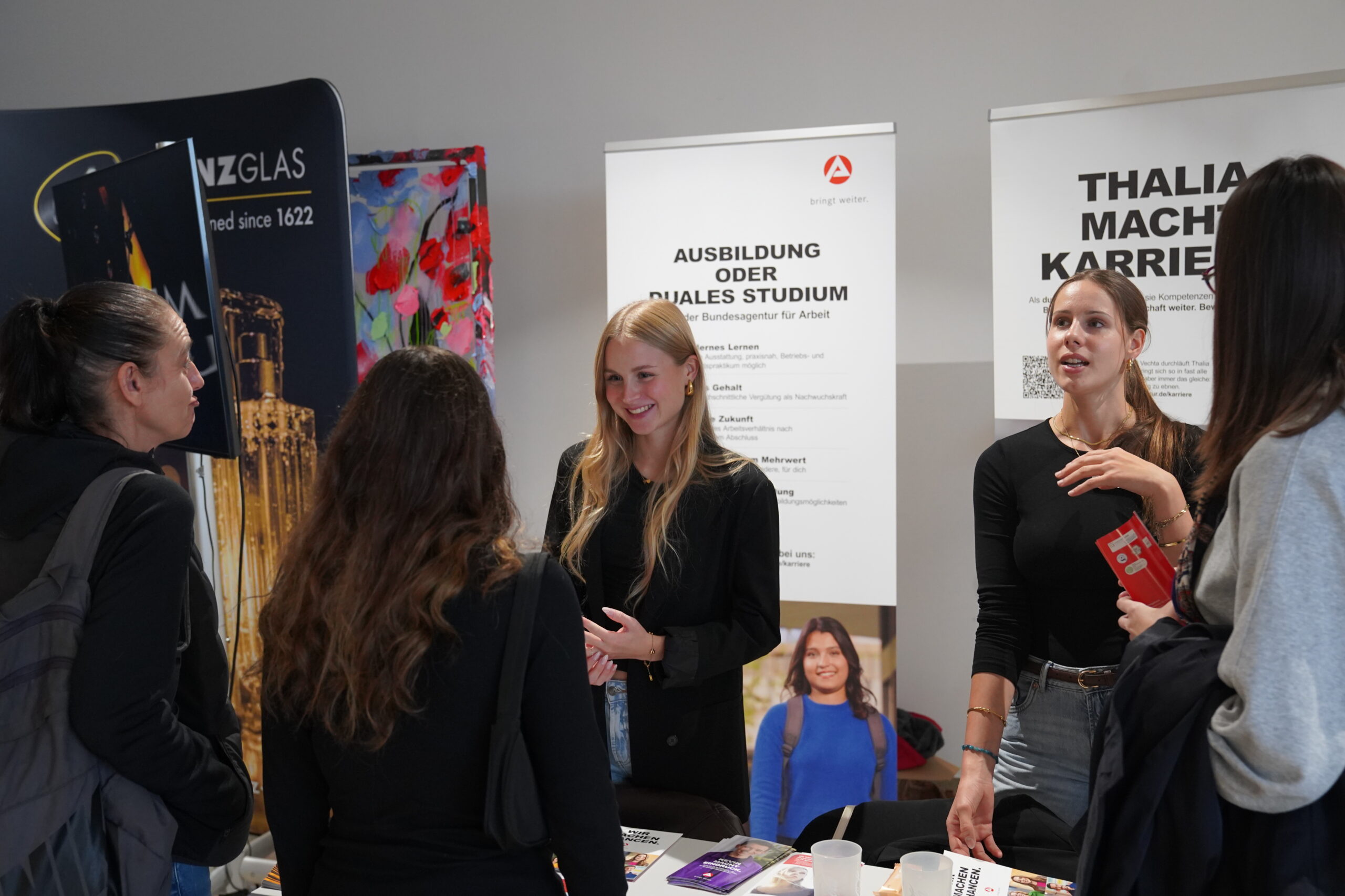 Vier junge Frauen unterhalten sich an einem Stand der Coburger Studienmesse, im Hintergrund sind Werbebanner zu sehen. at Hochschule Coburg