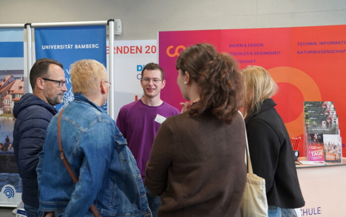 Ein Student in einem lila Hemd unterhält sich während der Coburger Studienmesse mit vier anderen Personen am Stand der Studienberatung einer Universität. at Hochschule Coburg