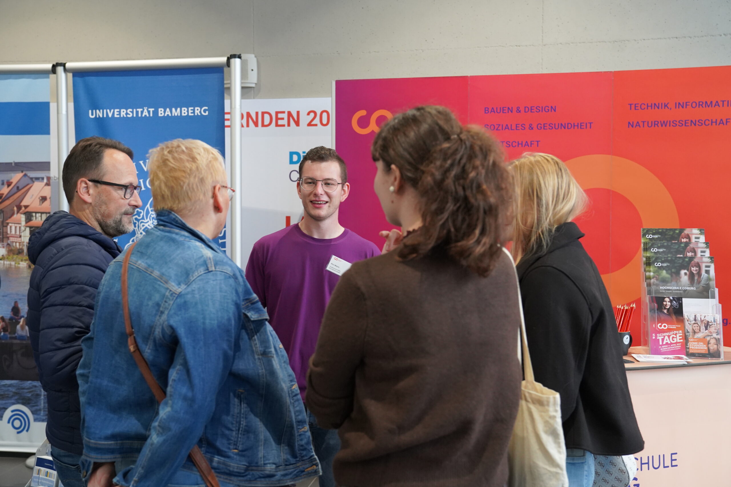 Ein Student in einem lila Hemd unterhält sich während der Coburger Studienmesse mit vier anderen Personen am Stand der Studienberatung einer Universität. at Hochschule Coburg