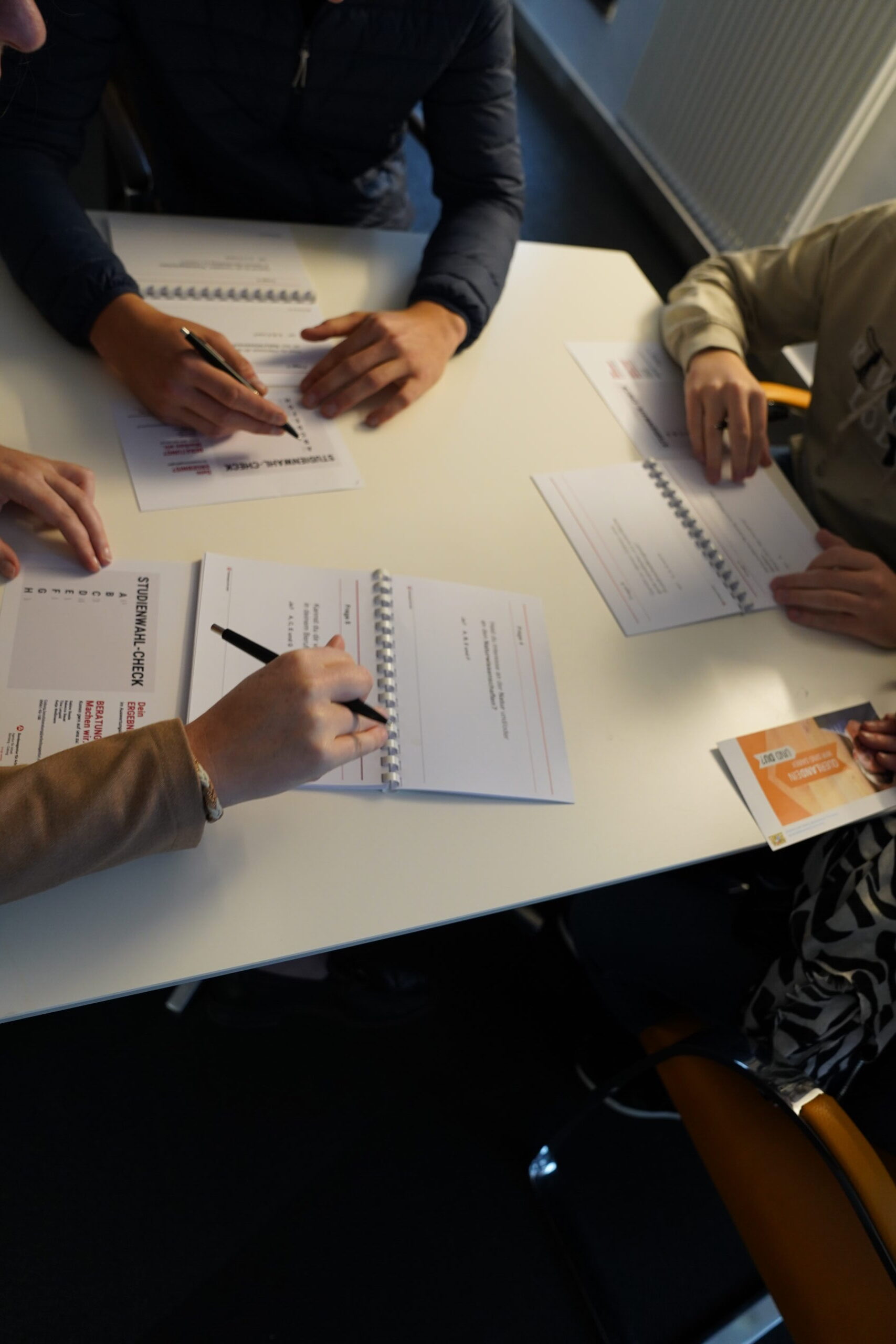 Vier Personen sitzen bei einer „Studienwahl-Check“-Veranstaltung im Rahmen der Coburger Studienmesse um einen weißen Tisch herum und machen sich Notizen in Spiralheften. at Hochschule Coburg