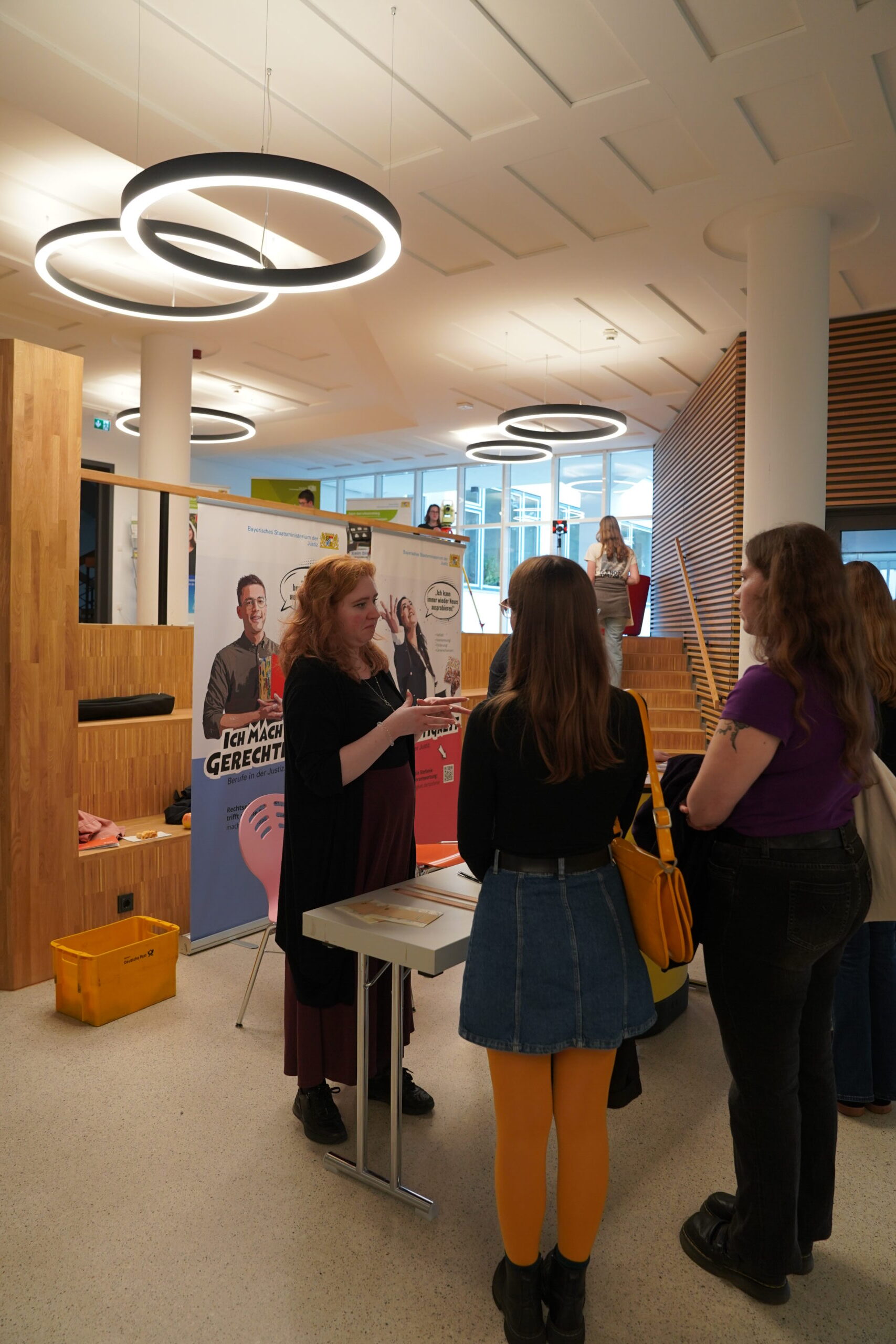 Eine Frau bietet an einem Informationsstand Studienberatung an und unterhält sich während der Coburger Studienmesse mit zwei Besuchern in der Lobby eines modernen Gebäudes. at Hochschule Coburg