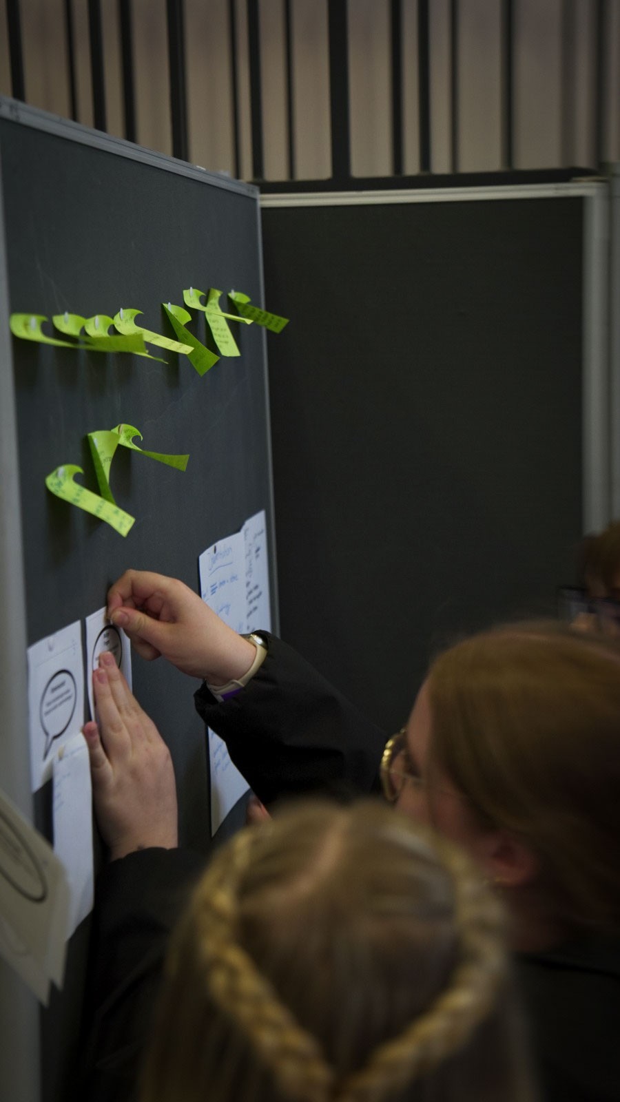 Eine Person klebt einen Haftnotizzettel an eine Anschlagtafel für die Veranstaltung „anCOmmen 2026“ an der Fakultät Design + Bauen, die mit Zetteln und Sprechblasensymbolen übersät ist. at Hochschule Coburg