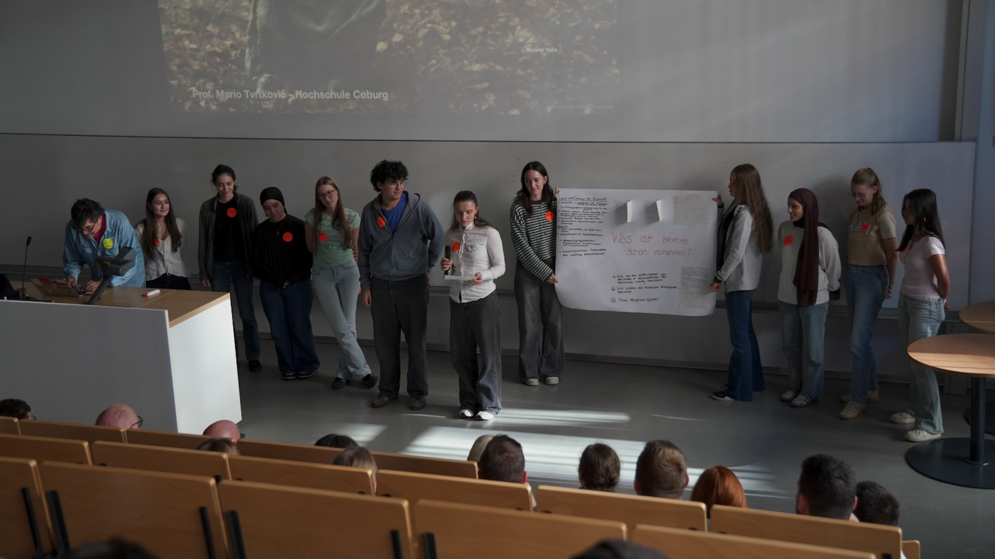 Eine Studentin hält ein Mikrofon in der Hand und spricht in einem Hörsaal zu den Zuhörern, während ihre Kommilitonen hinter ihr stehen; einer von ihnen hält ein großes Plakat für „aCOmmen 2026“ hoch. at Hochschule Coburg