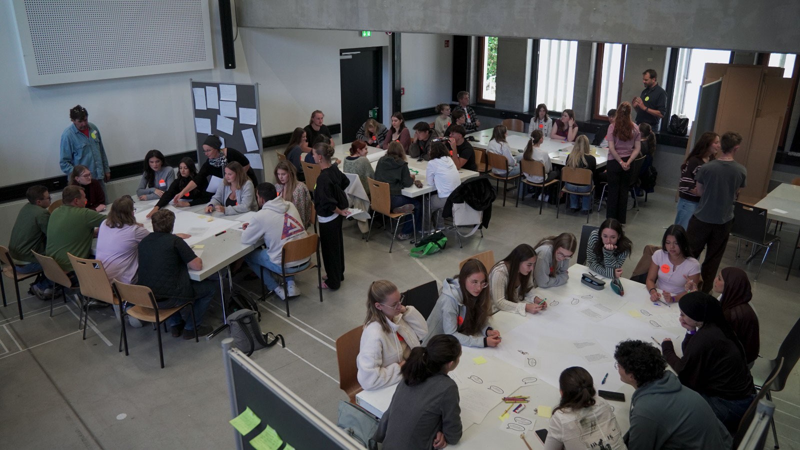 Erstsemester an der Fakultät Design + Bauen sitzen an Tischen in einem großen Raum und arbeiten in Gruppen an großen Papierbögen. at Hochschule Coburg