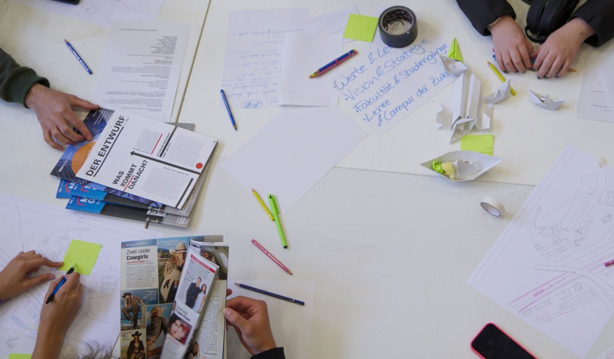 Vier Personen arbeiten an einem Tisch, der mit Broschüren, Skizzen und Papierbooten bedeckt ist, während der „anCOmmen 2026“-Einführungsveranstaltung für Studienanfänger der Fachrichtungen Design und Architektur. at Hochschule Coburg