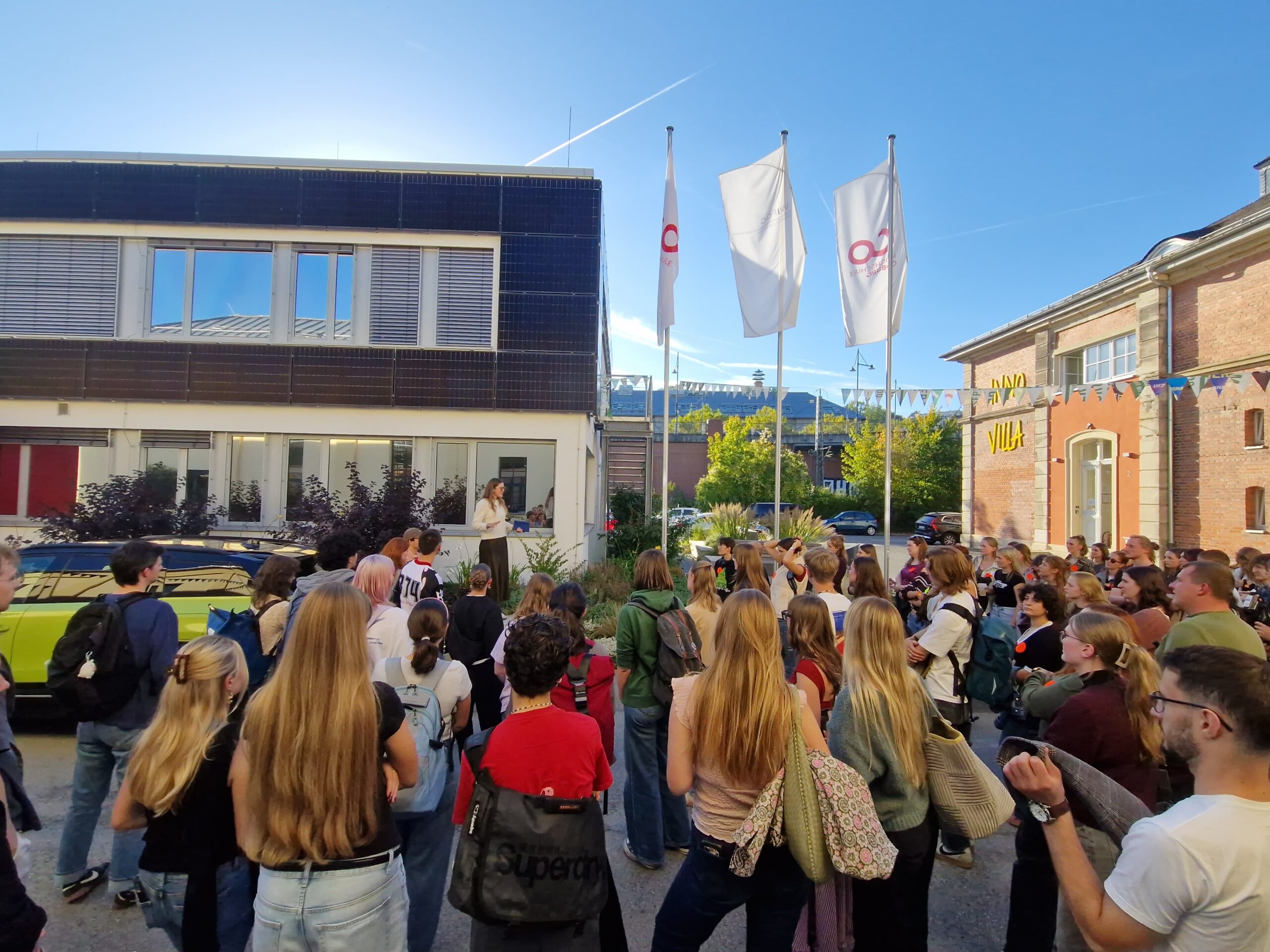 Eine Gruppe von Erstsemestern steht auf einem Innenhof und blickt zu einer Frau, die von einer erhöhten Bühne vor dem Gebäude „Design und Bauen“ spricht. at Hochschule Coburg