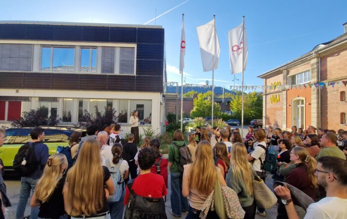 Eine Gruppe von Studierenden, die während ihres „Studium Coburg“ auf einem Innenhof steht und einer Frau zuhört, die vom Eingang eines Gebäudes aus spricht. at Hochschule Coburg