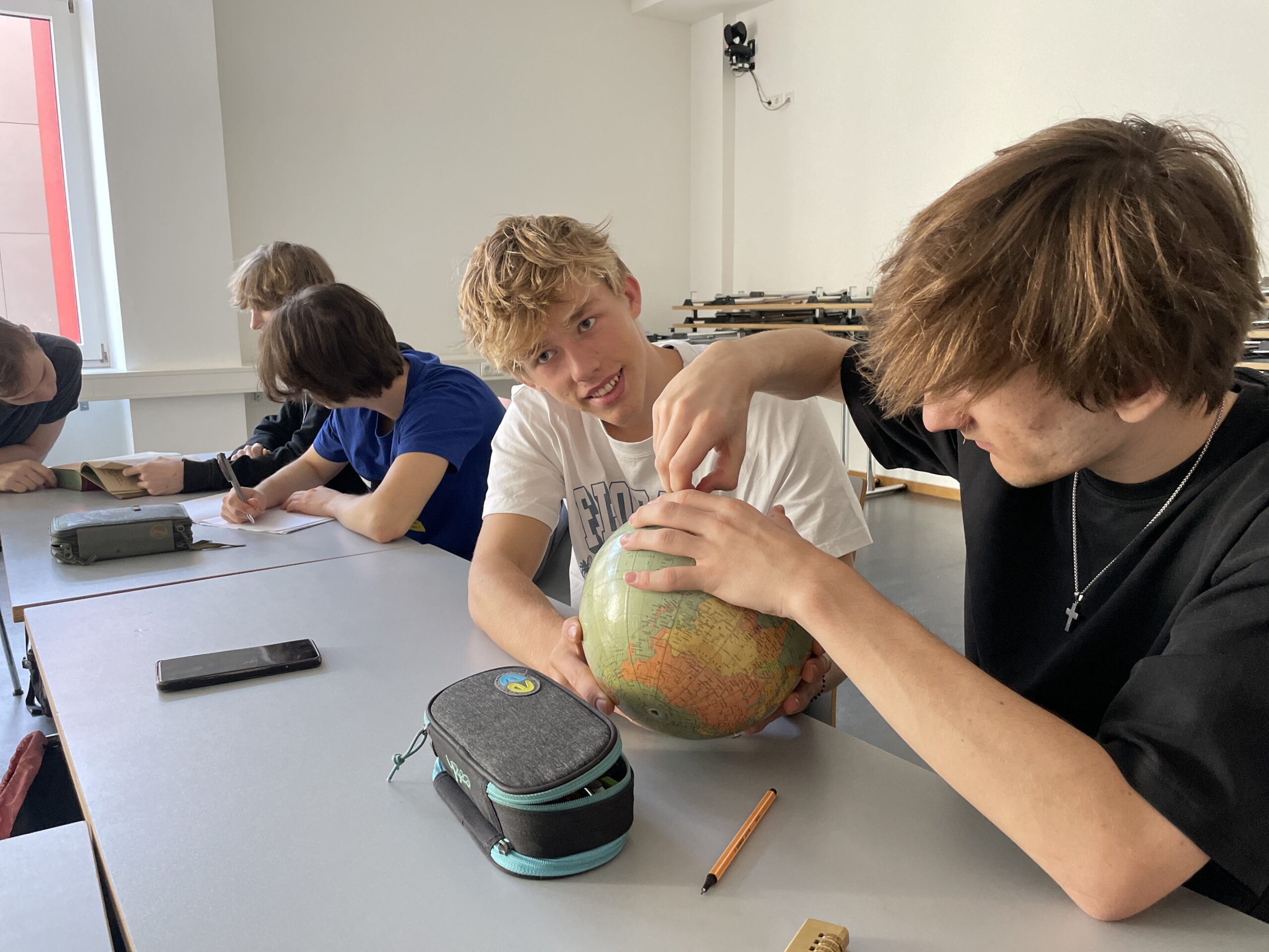 Zwei männliche MINT-Studenten betrachten gemeinsam einen Globus an einem Schreibtisch in einem Klassenzimmer, während andere Studenten in der Nähe lernen. at Hochschule Coburg