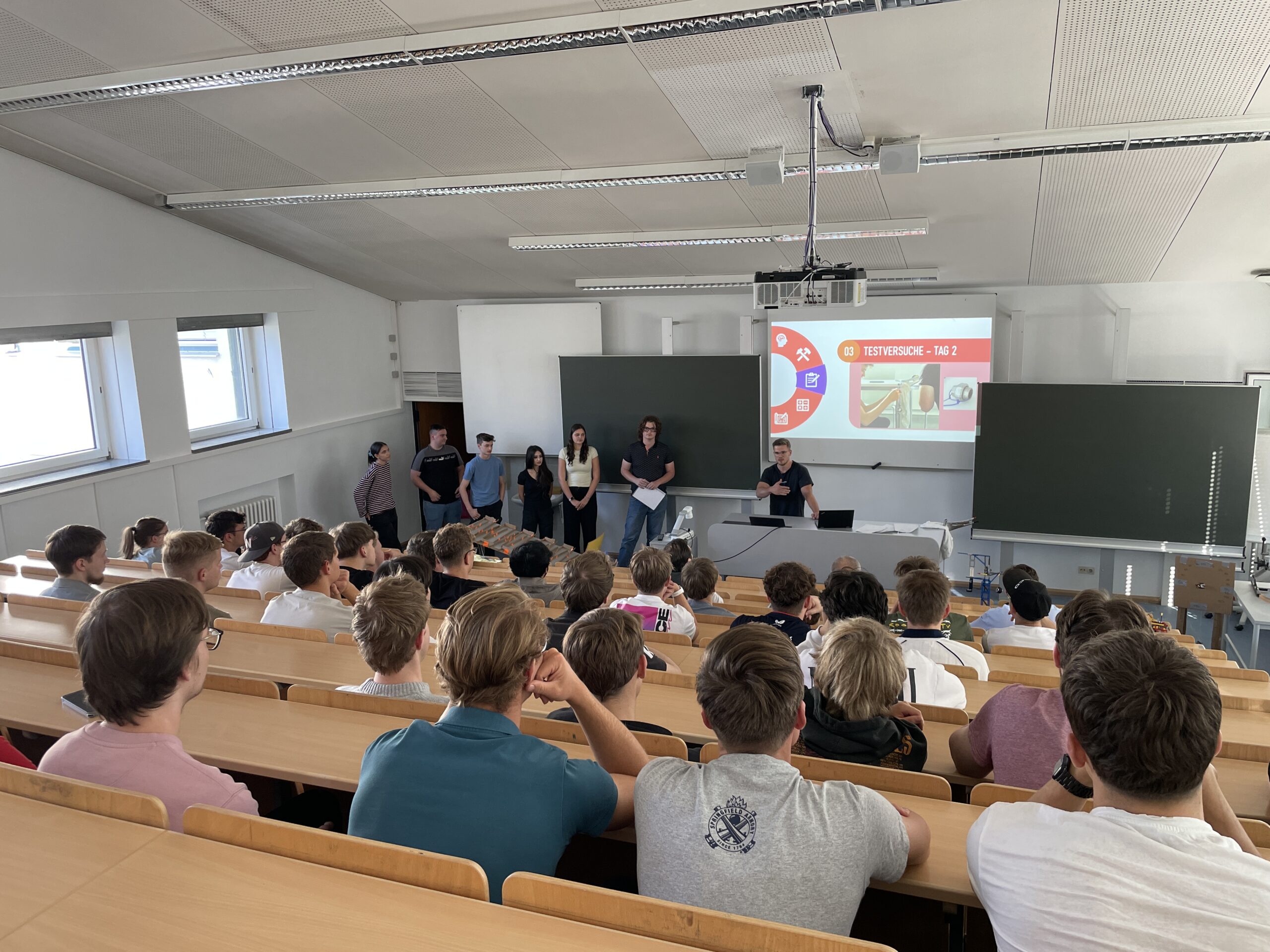 Eine Gruppe von MINT-Studierenden steht vor der Leinwand eines Hörsaals, auf der „Testversuche – Tag 2“ zu sehen ist, und blickt auf eine Klasse von Studierenden, die zu Beginn ihres Studiums im Hörsaal sitzen. at Hochschule Coburg