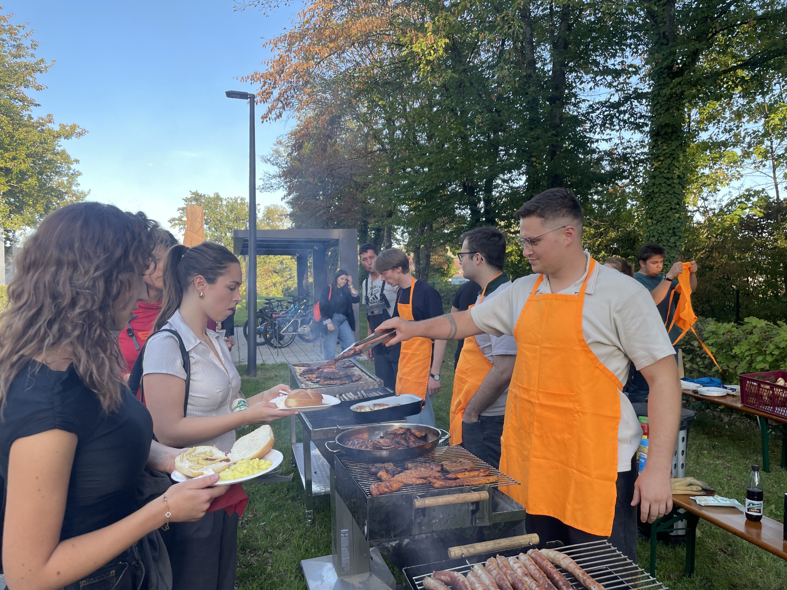 Ein junger Mann in einer orangefarbenen Schürze grillt bei einer Veranstaltung des MINT-Studiums Fleisch, während Erstsemester mit ihren Tellern Schlange stehen, um bedient zu werden. at Hochschule Coburg